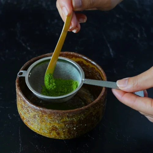 Japanese matcha being sifted through a fine stainless steel chakoshi into a bowl during tea preparation