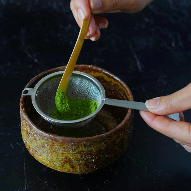Japanese matcha being sifted through a fine stainless steel chakoshi into a bowl during tea preparation
